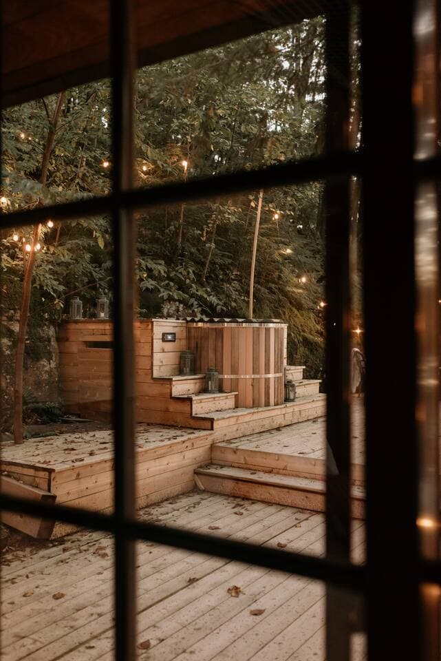 View through paned windows to the cedar hot tub and string-lit wooden deck in the forest.