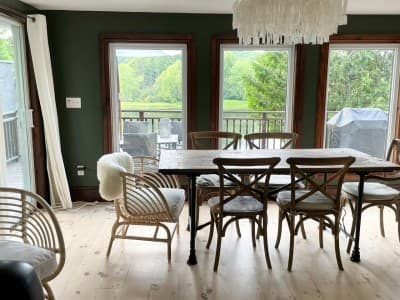 Dining area with rustic wood table, woven chairs, and big windows overlooking the deck and greenery.