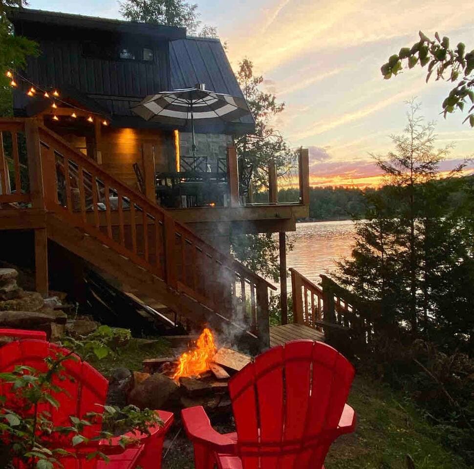Red Adirondack chairs circle a lakeside fire pit as the treehouse glows at sunset.