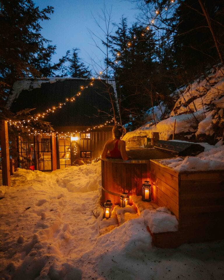 A guest soaks in the wood-fired hot tub on a snowy, string-lit winter evening.